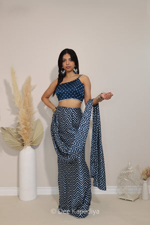 A woman modeling a navy blue and white chevron-patterned two-piece outfit consisting of a 1 shoulder bandhani print crop top and a matching pre-draped saree, standing in a studio with decorative dried grass in a vase and a small birdcage.