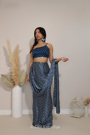 A woman modeling a navy blue and white patterned traditional outfit consisting of a 1 shoulder asymentrical bandhani print crop top, matching chevron patterned pre-draped saree , standing in a studio with decorative pampas grass and a birdcage.