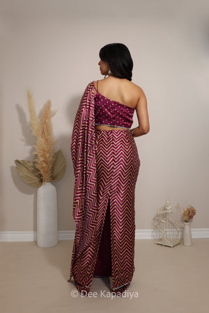 A woman stands with her back to the camera wearing a deep maroon and white chevron-patterned pre-draped saree with a matching embellished bandhani print blouse. The setting is a minimalist room with beige walls and floor, decorated with a tall vase of pampas grass on the left and small decorative wire cages on the right.