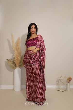 A woman modeling a traditional Indian saree with a deep magenta-pink and white chevron pattern pre-draped saree with matching bandhani print 1 shoulder asymetrical croptop, standing in a minimalist room with beige walls and decorative dried grass in a white vase.