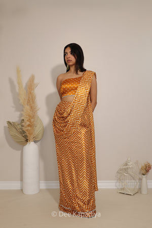 A woman modeling a golden yellow and orange bandhani print 1 shoulder blouse croptop with chevron-patterned spre-draped aree with a matching sleeveless blouse, standing in a studio with decorative dried grass and a small birdcage on a beige floor against a cream wall.