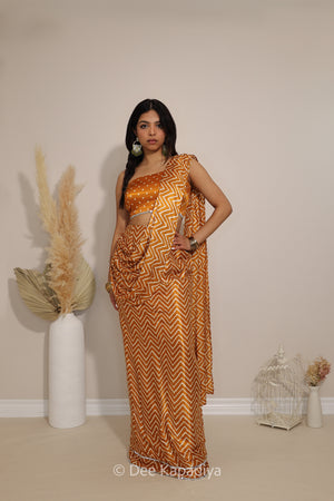 A woman modeling a metallic gold and yellow bandhani print one shoulder blouse croptop and chevron-patterned pre-draped saree with a matching sleeveless blouse, standing in a studio with decorative dried grass and a birdcage.