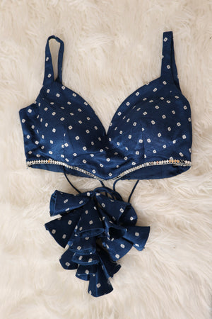 A indigo satin bralette croptop with white bandhani print and a real mirror-studded underband, displayed flat on a white faux fur background with matching ruffled bow accessories below.