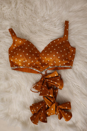 An orange satin bralette croptop with white bandhani print and a real mirror-studded underband, displayed flat on a white faux fur background with matching ruffled bow accessories below.