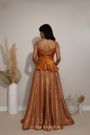 A woman stands with her back to the camera wearing a sleeveless haldi yellow and white chevron patterned lehenga with a bandhani print peplum waist and a long chevron-patterned skirt. The photo is taken in a studio setting with decorative dried grass in a white vase and a small white birdcage on a beige floor against a plain beige wall.