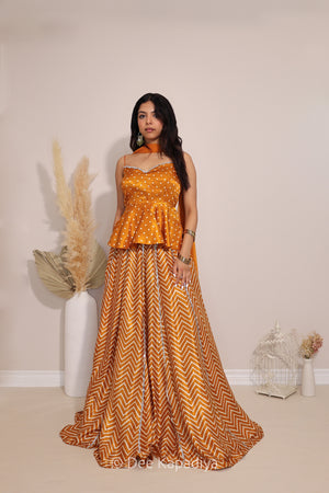A woman models a traditional Indian outfit consisting of a mustard yellow and white bandhani printed patterned peplum top and matching chevron printed skirt with silver zari embroidery, set against a plain beige wall with decorative dried pampas grass.
