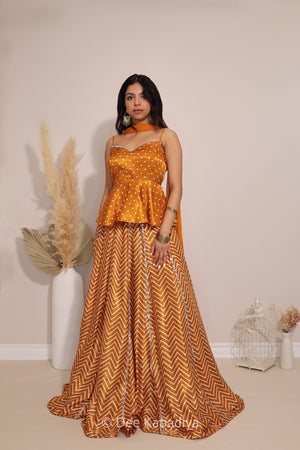A woman models a two-piece traditional Indian outfit consisting of a mustard yellow top with bandhani print and a matching  chevron print skirt with a haldi yellow and white chevron pattern, set against a neutral beige background.