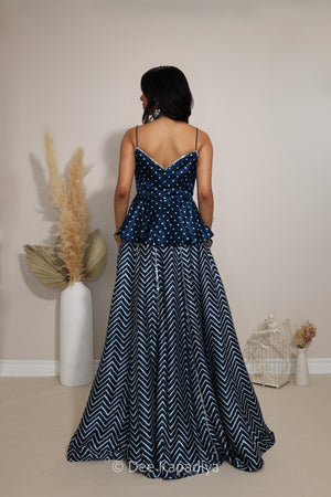 A woman viewed from behind wearing a sleeveless navy lehenga with a white bandhani print peplum bodice and a navy-and-white chevron patterned skirt, standing in a studio with decorative dried grass and a birdcage.