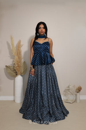 A woman modeling a navy blue formal gown with a bandhani printed bodice and a chevron-patterned skirt, standing in a studio setting with decorative dried grass and a birdcage.