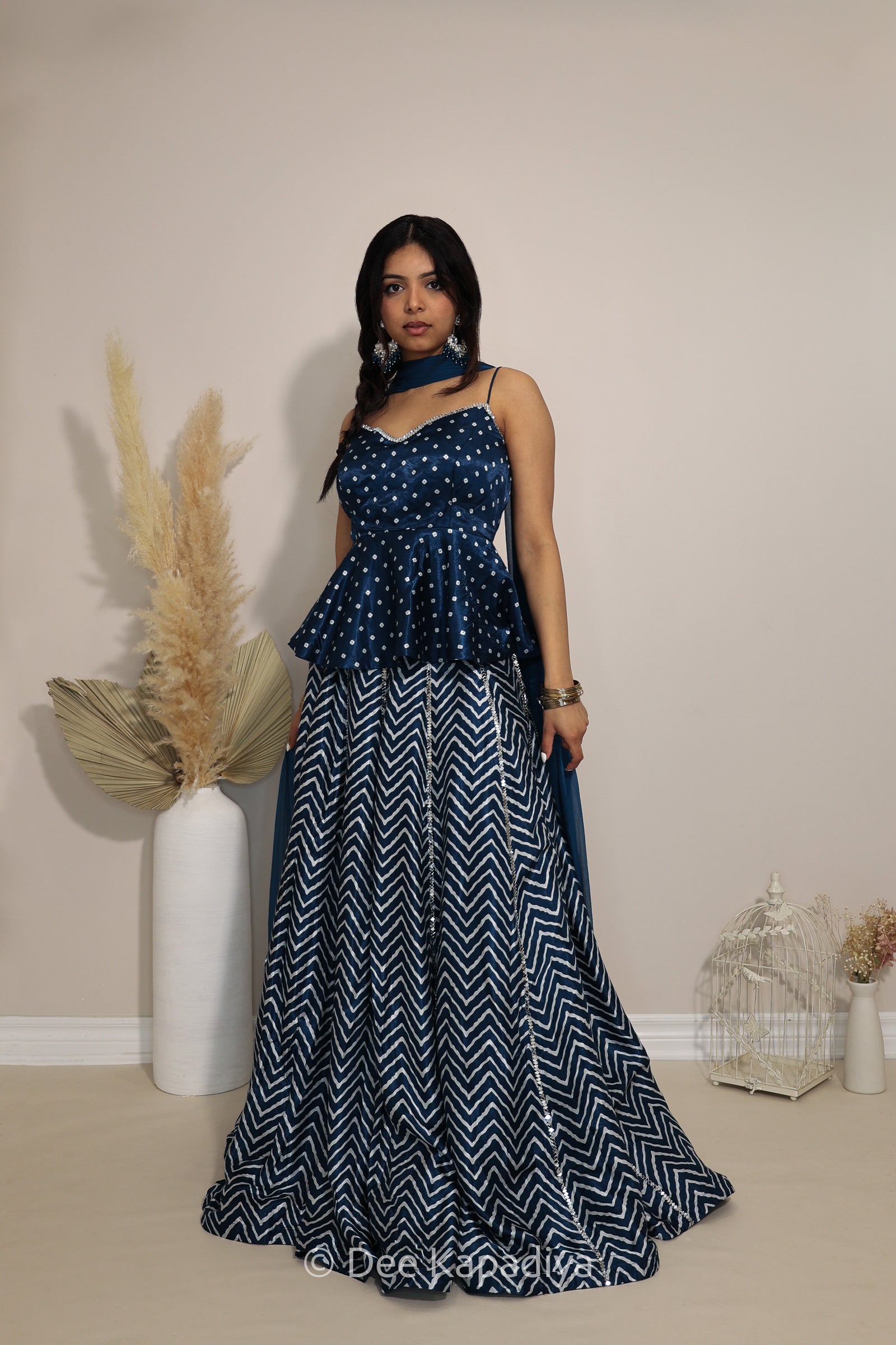 A woman modeling a navy blue and white printed traditional Indian lehenga in chevron print with a top, peplum waist in bandhani print, and a matching navy blue scarf, standing in a studio setting with decorative pampas grass.