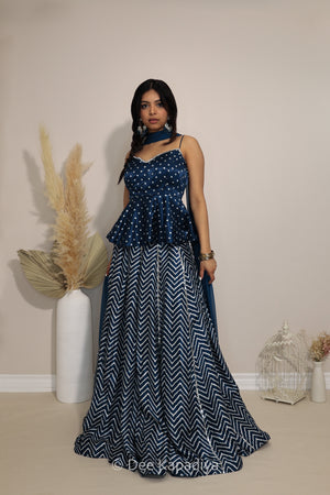 A woman modeling a navy blue and white printed traditional Indian lehenga in chevron print with a top, peplum waist in bandhani print, and a matching navy blue scarf, standing in a studio setting with decorative pampas grass.