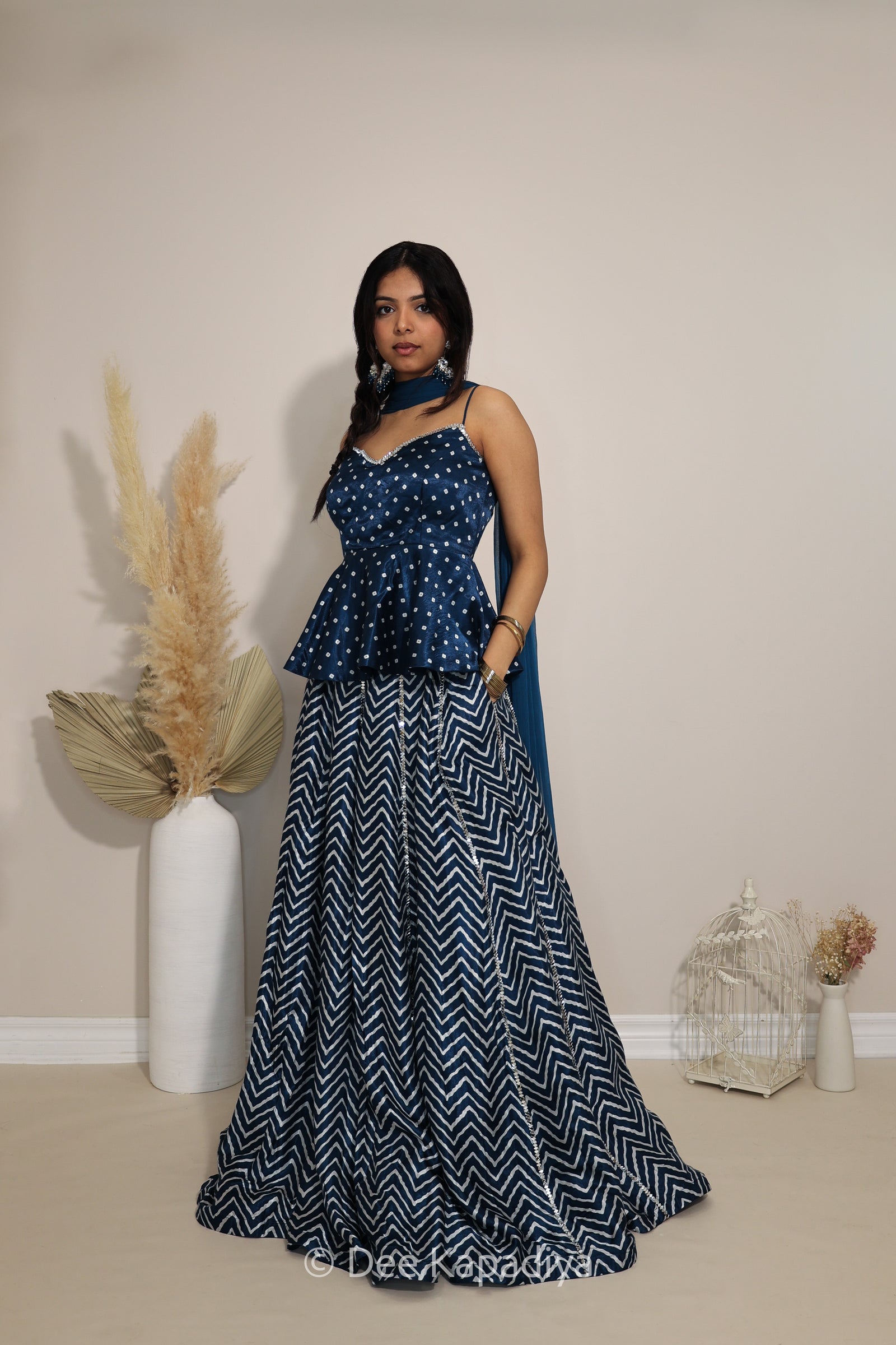 A woman modeling a navy blue and white printed traditional Indian lehenga in chevron print with a top, peplum waist in bandhani print, and a matching navy blue scarf, standing in a studio setting with decorative pampas grass.
