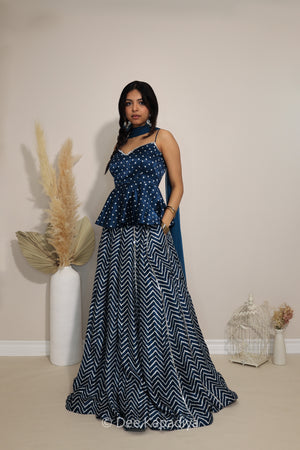A woman modeling a indigo blue traditional lehenga with a bandhani print bodice and a navy-and-white chevron-patterned skirt-lehenga, standing in a studio setting with decorative dried grass in a white vase.