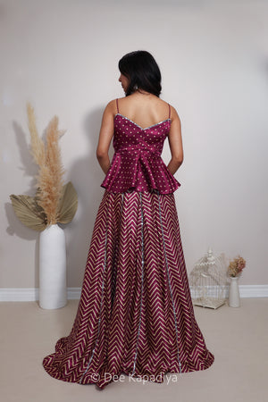 A woman viewed from behind wearing a floor-length lehenga with a deep V-back, embellished burgundy bodice with silver bandhani print and a matching chevron-patterned skirt. She is standing in a studio setting with decorative dried grass in a white vase and a small white birdcage on a beige floor against a light gray wall.