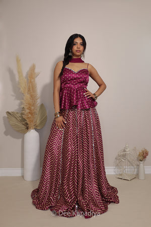 A woman models a two-piece magenta and burgundy ethnic outfit consisting of a sleeveless bandhani print peplum top and a matching chevron print lehenga skirt, standing in a studio with decorative pampas grass.