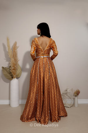 A woman stands with her back to the camera wearing a floor-length orange and gold chevron patterned lehenga with a deep V-neckline and long sleeves ajrakh print blouse. The dress features a geometric chevron-like pattern.