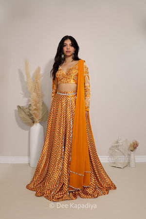 A woman models a traditional Indian outfit consisting of a mustard yellow and orange chevron-patterned lehenga skirt, matching embroidered ajrakh print crop top, and a solid orange dupatta, set against a neutral beige wall with decorative dried grass in a white vase.