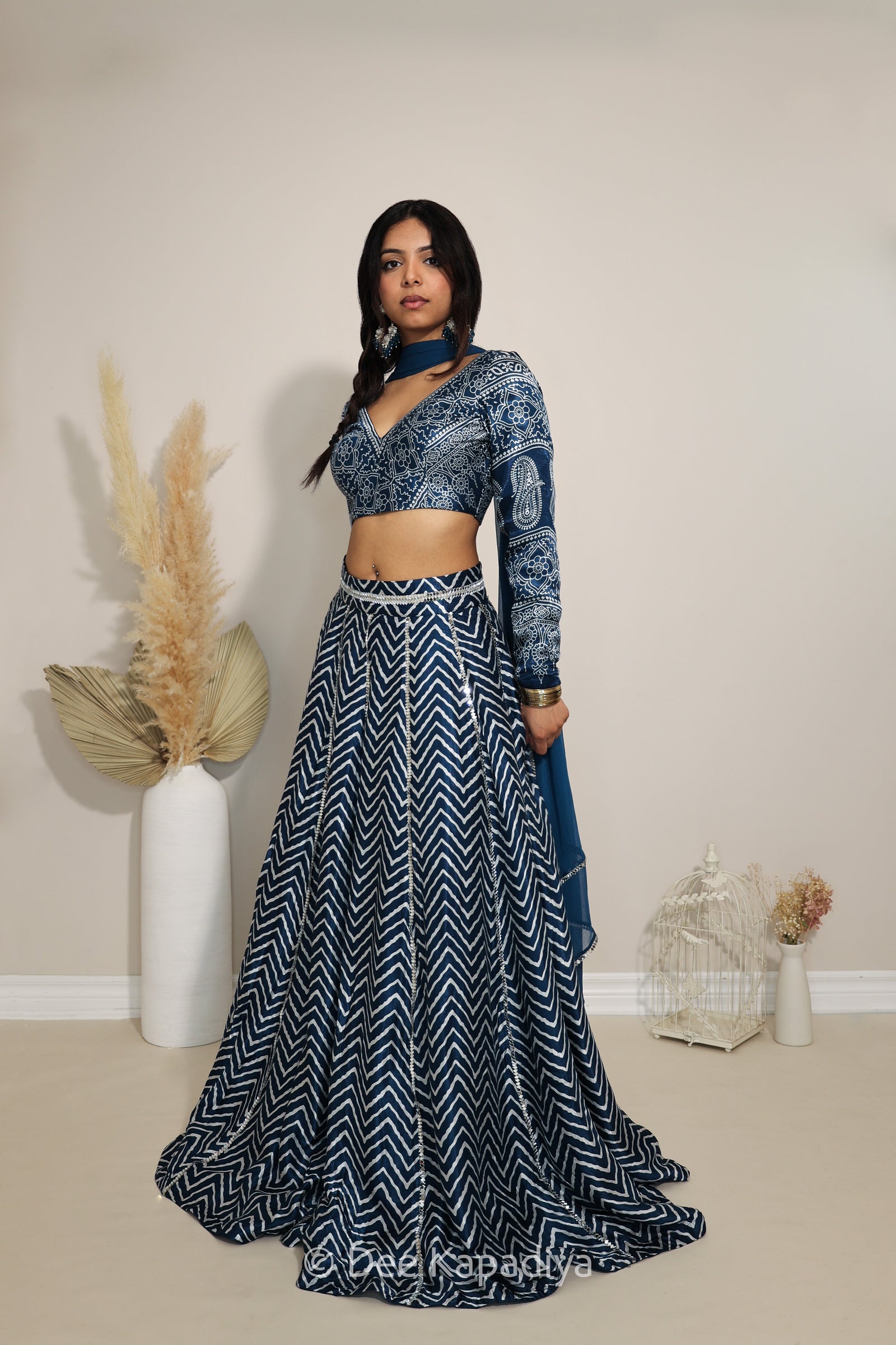 A woman models a navy blue and white patterned Indian ethnic outfit consisting of a long-sleeve ajrakh print crop top, a matching flared chevron printed skirt, and a draped scarf, standing in a studio with decorative dried grass in a vase.