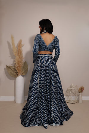 A woman stands with her back to the camera wearing a dark blue and white patterned two-piece outfit with a long-sleeve ajrakh print crop top and a flared chevron print skirt. The setting is a neutral indoor studio with decorative dried grass in a white vase and a small white birdcage.