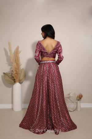 A woman stands with her back to the camera wearing a deep maroon and gold chevron-patterned two-piece outfit, consisting of a long-sleeve ajrakh print blouse and a matching flared skirt. The setting is a neutral indoor studio with decorative dried pampas grass in a white vase.