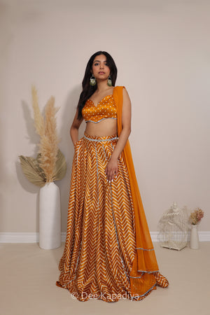 A woman models a traditional Indian outfit consisting of a mustard yellow and gold chevron-patterned lehenga skirt, a matching crop top with bandhani print, and a solid orange dupatta, posed in front of a neutral beige wall with decorative pampas grass.