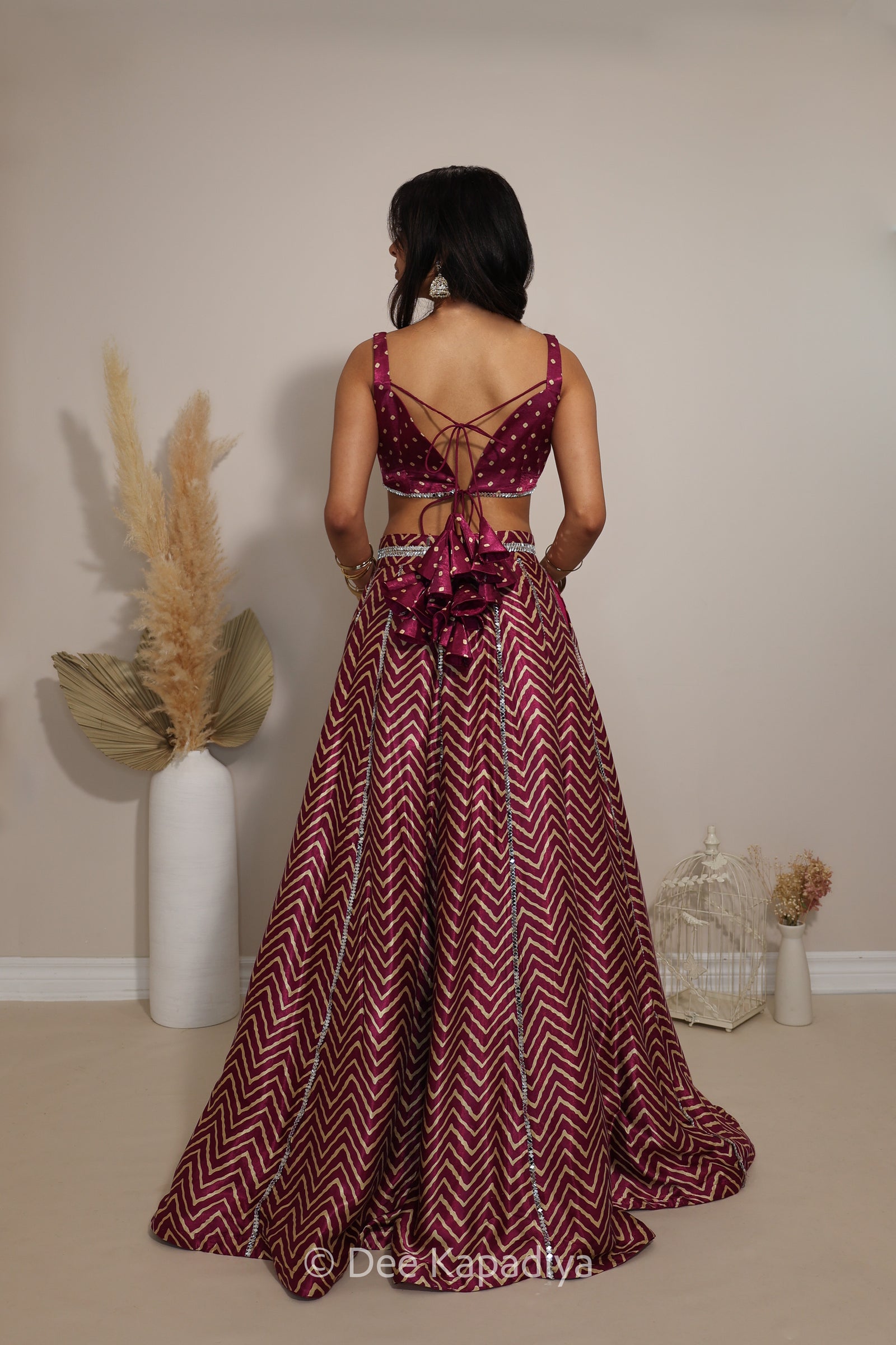 A woman stands with her back to the camera wearing a floor-length maroon and burgundy chevron-patterned lehenga with a lace-up open back and a large bow at the waist in bandhani print. The photo is taken indoors against a plain beige wall with decorative dried grass in a white vase and a small white birdcage.