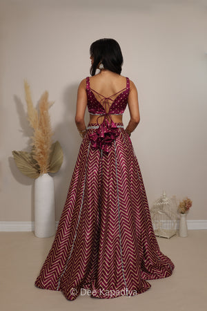 A woman stands with her back to the camera wearing a floor-length maroon and burgundy chevron-patterned lehenga with a lace-up open back and a large bow at the waist in bandhani print. The photo is taken indoors against a plain beige wall with decorative dried grass in a white vase and a small white birdcage.