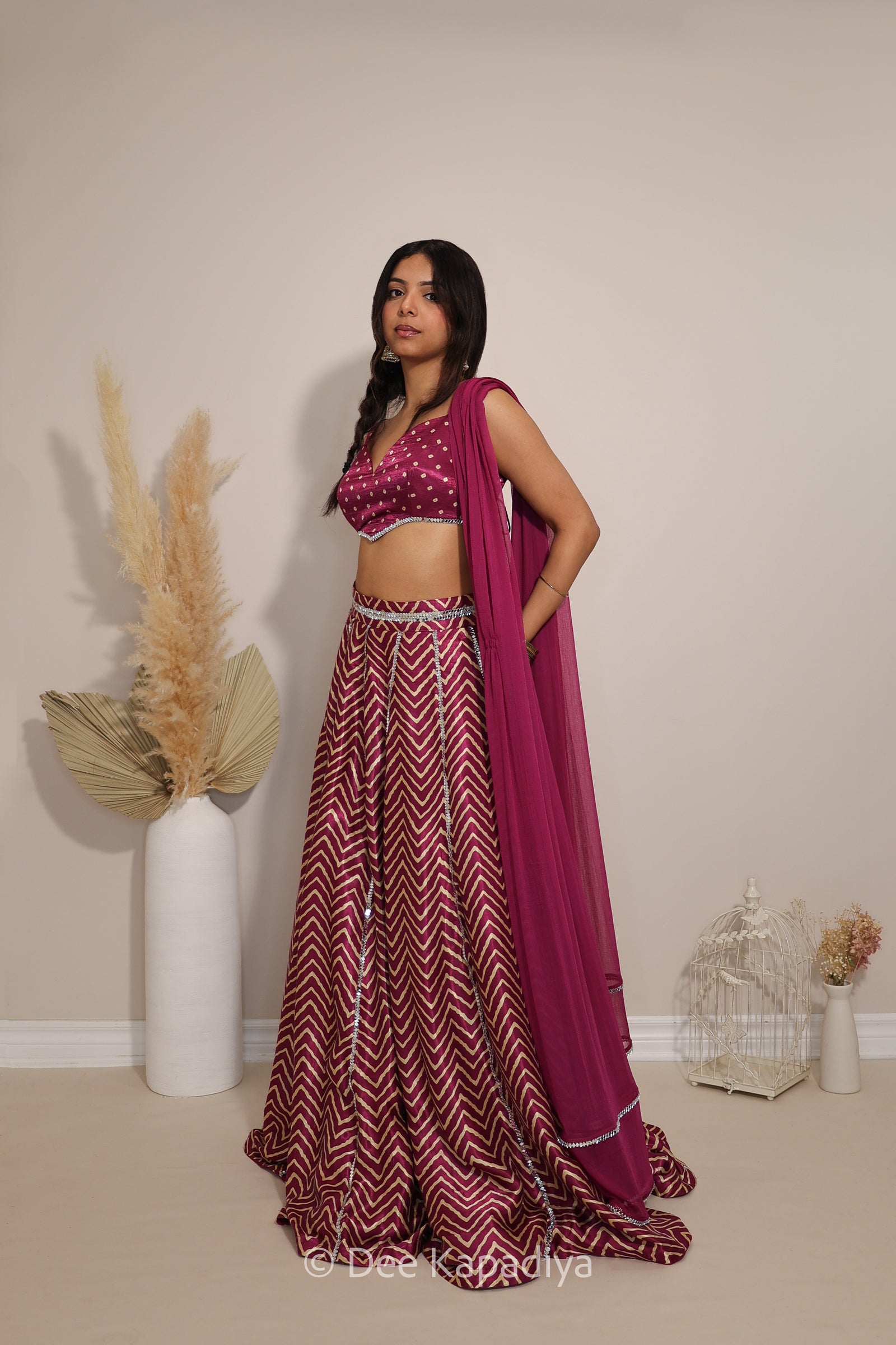 A woman stands with her back to the camera wearing a floor-length maroon and burgundy chevron-patterned lehenga with a lace-up open back and a large bow at the waist in bandhani print. The photo is taken indoors against a plain beige wall with decorative dried grass in a white vase and a small white birdcage.